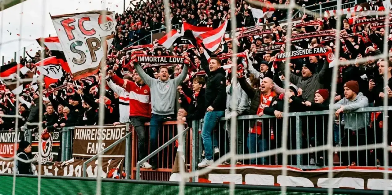 Aficionados del fútbol alemán en las gradas de un estadio de la 2. Bundesliga celebrando un gol con bufandas y banderas del equipo
